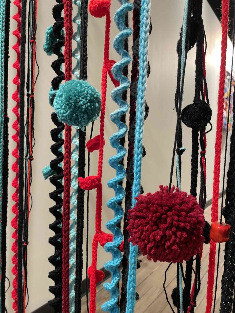 A close-up view of colorful yarn strands hanging vertically. The strands are in black, red, and various shades of blue. Some strands are straight while others are twisted or curled. There are also pom-poms in red and light blue attached to some of the strands. The overall effect is of a vibrant, textured textile art piece or decoration