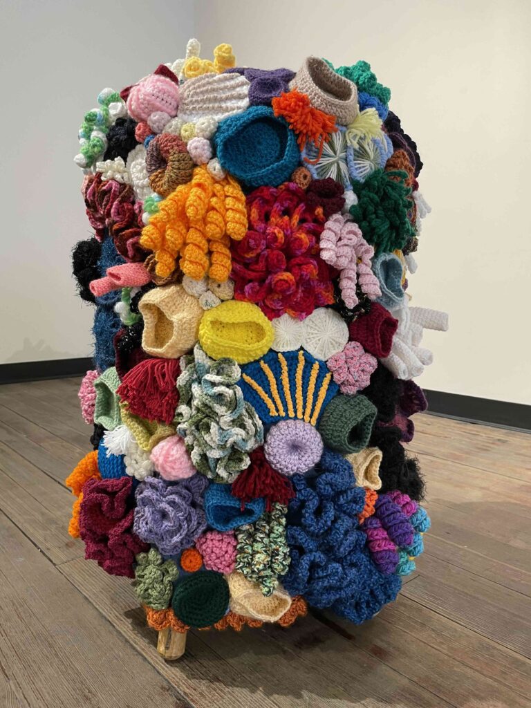 A vibrant, three-dimensional textile art sculpture sitting on a wooden floor. The sculpture is composed of a densely packed array of crocheted and knitted elements in a wide variety of colors, textures, and shapes. It includes flowers, spirals, pom-poms, and abstract forms, creating a riot of colors including reds, blues, yellows, greens, purples, and oranges. The overall shape is roughly cylindrical or column-like, standing about waist-high.