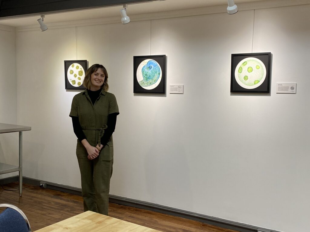 Samantha stands in a green jumpsuit and black long-sleeve shirt smiling in Lake City art gallery. Behind her on a white wall are three framed circular artworks of her microscopic illustrations - one featuring yellow-green organisms, one in blue tones, and one with green dots on a textured white background. Track lighting illuminates the artwork