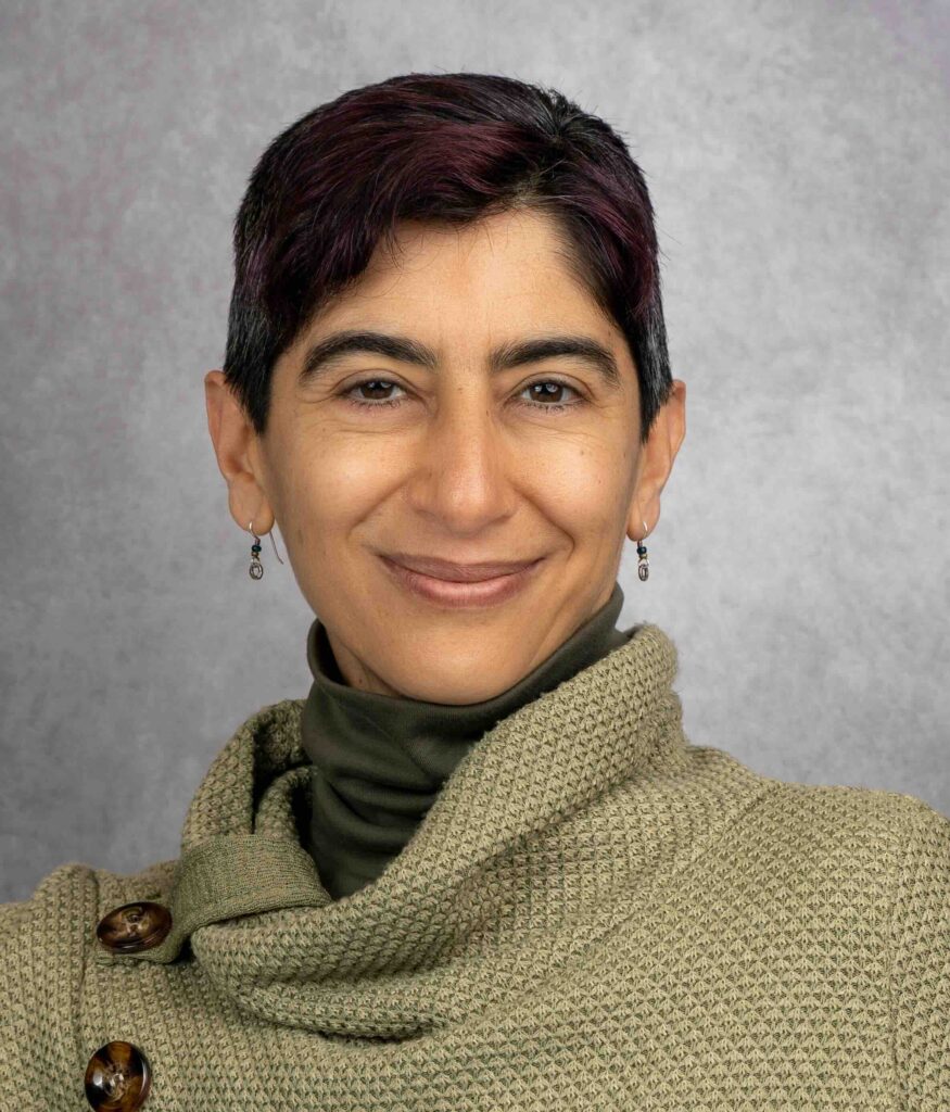 Professional headshot of Sharon Mansur smiling warmly at the camera. She has short dark hair with subtle burgundy highlights, and wears small earrings. She's dressed in an olive green turtleneck under a knit sweater in sage green with large brown buttons. The photo is taken against a neutral gray background