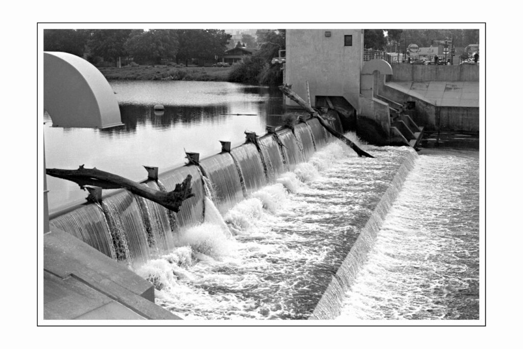 A black and white photograph of a dam or weir structure spanning a river. Water cascades over the concrete barrier creating a waterfall effect. The structure has several metal or wooden supports protruding along its length. The scene includes a curved architectural element on the left side and a building structure on the right, with calm water visible in the background.