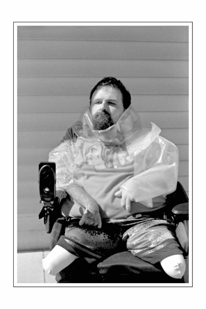 A black and white photograph of a person in a wheelchair wearing protective gear including what appears to be a waterproof poncho or protective suit. The individual has a beard and is seated against a background of horizontal siding or paneling. They appear to be wearing shorts and the image shows their prosthetic or artificial legs.