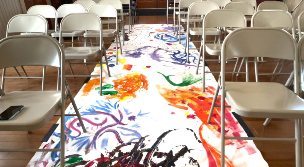 An aisle covered with a white canvas displaying colorful collaborative artwork with vibrant paint splashes in blues, oranges, purples, and greens. The table is surrounded by empty white folding chairs in what appears to be a community room or workshop space with a wooden floor.