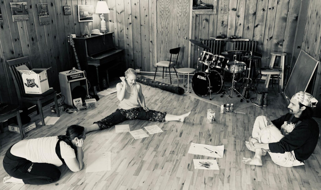 AA black and white photograph showing three people in a music or rehearsal space with wooden paneled walls. One person sits on the floor in a split position looking at papers, while two others sit nearby. The room contains musical equipment including a drum set, piano, and an amplifier.