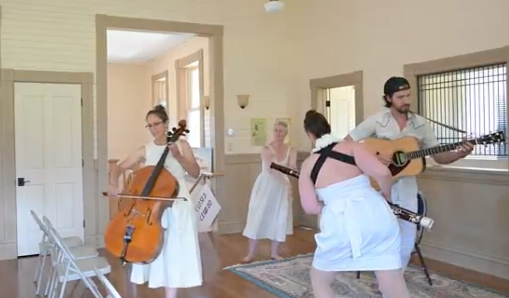 Four musicians performing in a bright interior space with cream-colored walls. A woman in a white dress plays cello, another musician plays a wind instrument, and a man with a backward cap plays acoustic guitar. All performers are dressed in white or light-colored clothing in what appears to be a small concert or recording session.