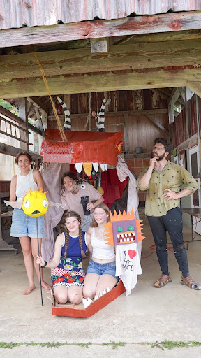 A group of people posing with handmade puppets and props inside what appears to be a rustic barn or wooden structure. The group includes both adults and children holding colorful puppet creations, including what looks like a boat or vessel with decorative elements. They are smiling at the camera, showcasing their theatrical crafts in the wooden interior space.