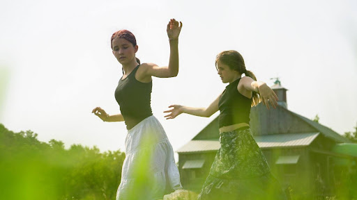 Two young performers in dance poses in a grassy field. Both are wearing dark tops with light-colored skirts or pants. They appear to be in mid-movement with their arms gracefully positioned. In the background, there's a building with a distinctive roof visible through the sunlit haze.