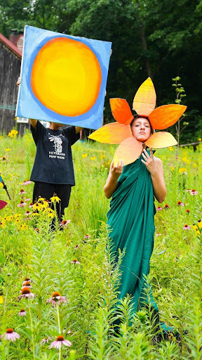 A photograph taken in a flower-filled meadow showing two people performing outdoors. One person wearing a black shirt is holding up a large square painting or prop of a bright yellow sun with blue background. Next to them stands a person dressed in a green wrap or dress wearing a large orange and yellow flower headdress or costume piece. They are surrounded by tall wildflowers and greenery in what appears to be a performance or artistic presentation in a natural setting.