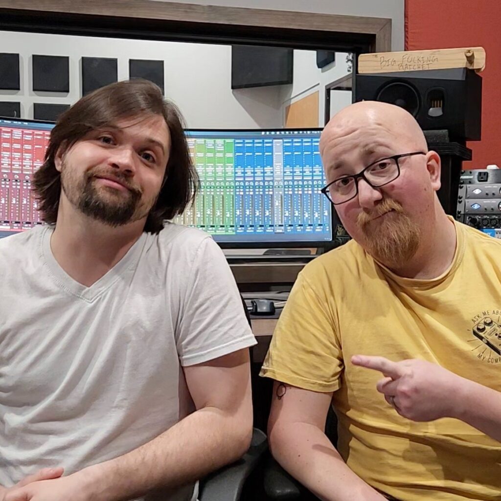Two men sitting together in a recording studio. On the left is Oliver Books, wearing a white v-neck t-shirt, with shoulder-length brown hair and a beard. On the right is Alex Oberg, wearing a yellow t-shirt and glasses, who is bald with a reddish beard and pointing at the camera. Behind them is professional audio equipment including mixing consoles with colorful channel displays, monitors, and studio gear. A wooden sign reading 'Pro Focusing Hatchet' is visible on the equipment rack.