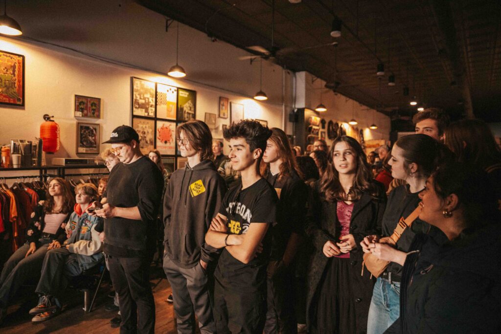 "A crowded Treedome Vintage and Vinyl. A diverse group of young people are standing and watching something off-camera, with expressions of interest and engagement. The audience includes teenagers and young adults wearing casual clothing like hoodies, t-shirts, and jeans. The venue has exposed black ceiling beams, pendant lighting, and walls decorated with framed album covers and music posters. Clothing racks are visible on the left side, and the warm lighting creates an intimate, underground music scene atmosphere. The crowd appears to be attending a live performance or music event.