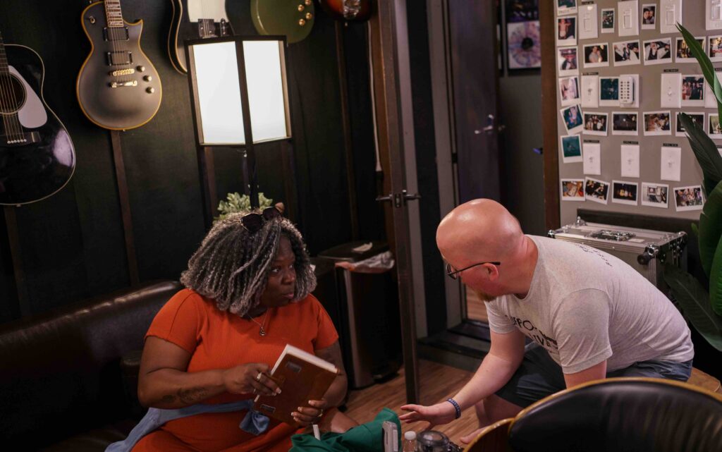 LaSonya Natividad with gray locs wearing an orange top sits on a dark leather couch holding a book, while a bald white man in glasses and a light-colored t-shirt leans over from his position on what appears to be a wooden chair or stool, reaching toward a green item on a coffee table. The setting is a cozy recording studio or music room with guitars mounted on the dark walls, a music stand with sheet music, and a wall covered in photographs and memorabilia. The warm lighting and intimate atmosphere suggest this is a creative workspace or interview setting."