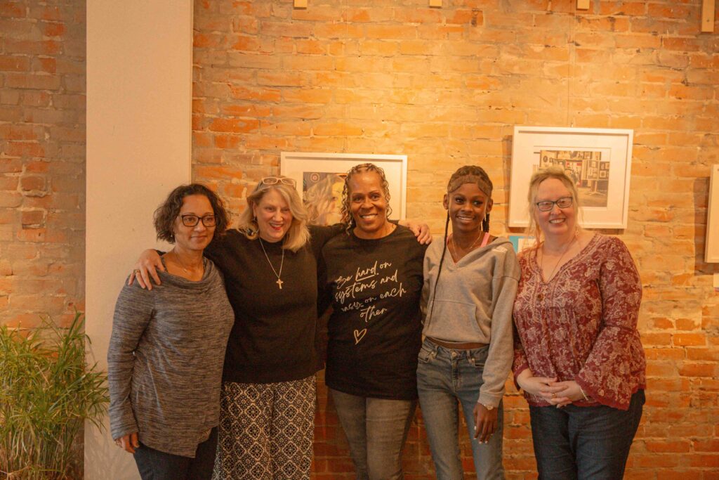 Five women standing together with their arms around each other in a warm group photo against an exposed brick wall. From left to right: a woman with dark curly hair and glasses in a gray striped top, a blonde woman in black with a cross necklace, a woman in a dark sweatshirt with cursive text, a young woman in a gray hoodie and jeans, and a woman with glasses in a red patterned blouse. Framed artwork is visible on the brick wall behind them, and there's a plant visible in the lower left corner.