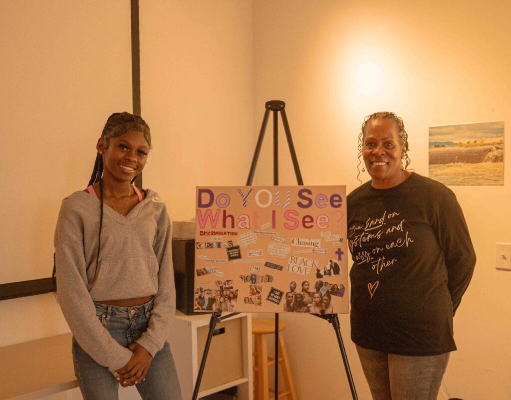 Two women standing beside a colorful poster on an easel titled "Do You See What I See?" The woman on the left wears a gray hoodie and jeans with her hair in braids, while the woman on the right wears a dark sweatshirt with cursive text and has curly hair. The poster features various images, text about discrimination, and phrases including "BLACK LOVE" and "Chasing" among other community-focused content.