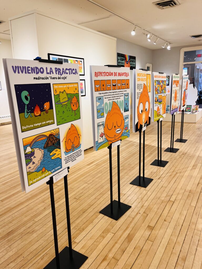 Gallery space with wooden floors showing a row of educational poster displays on black stands. The posters feature colorful cartoon characters and are titled in Spanish including "VIVIENDO LA PRACTICA" and "REPETICION DE MANTRA" with bright orange flame characters and other cartoon figures.