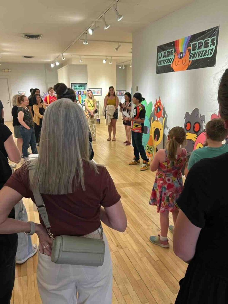 Crowded gallery opening with visitors of various ages standing and sitting on wooden floors. Large colorful cartoon character cutouts are mounted on the white walls in the background and a banner reading "U ARE UNIVERSE/ERES EL UNIVERSO" hangs from the ceilin
