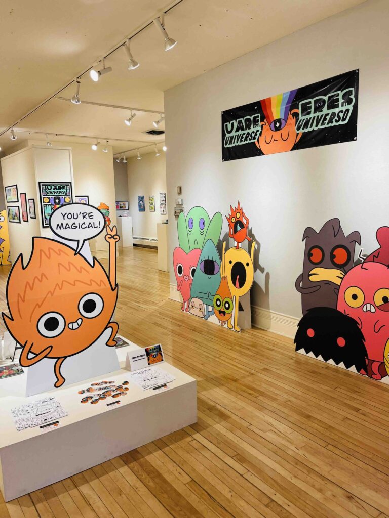 Photo provided by grant recipient Gallery interior showing large cartoon character cutouts mounted on white walls including a prominent orange character saying "YOU'RE MAGICAL" in a speech bubble. A white display table in the foreground contains printed materials and the exhibition banner is visible overhead.