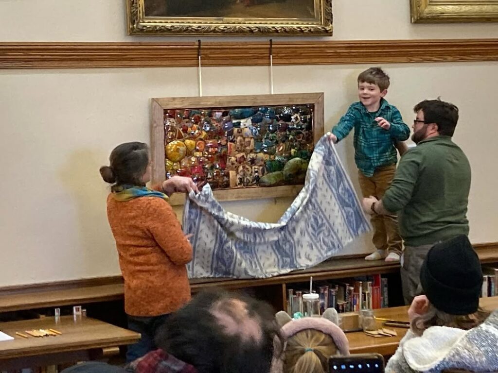 The Pauli Family holding up a decorative fabric or blanket in front of a wall-mounted display case containing Rachel Pauli's gourd mosaic unveiling.