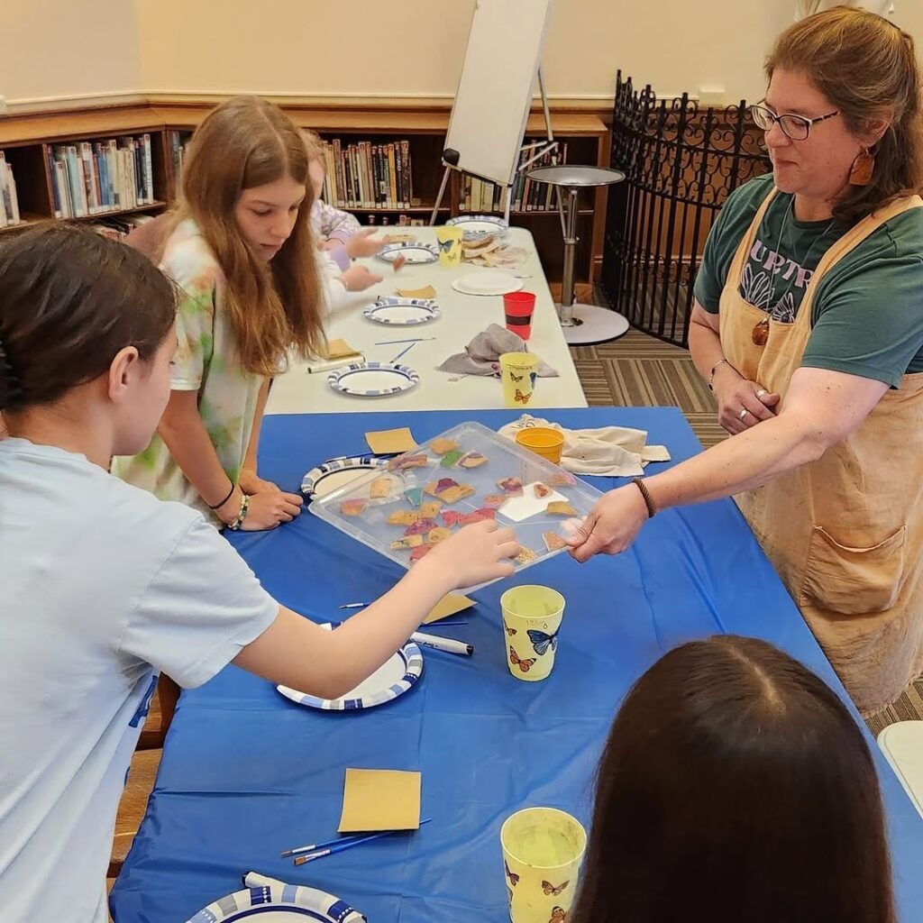 Children and an instructor working on an art project at a table with mosaic pieces, painting plates decorated with blue and white patterns during Rachel Pauli's gourd project workshop
